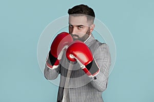 Serious young businessman in boxing gloves ready to fight against blue background