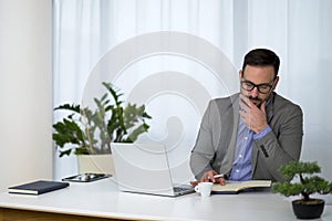 Serious young businessman with short stylish beard and glasses sitting at office and taking notes