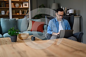 Serious young businessman wearing eyeglasses sending business e-mails over laptop while sitting on armchair. Confident male