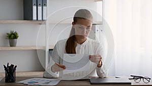 Serious young businesswoman opening envelope and reading letter at workplace in office