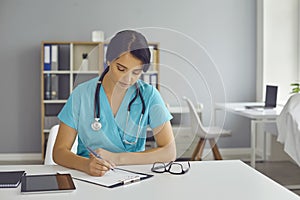 Serious young doctor sitting at desk and completing information in patient medical history