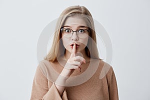 Serious young female with blonde straight hair, wearing eyeglasses, dressed causally holding finger at her lips asking