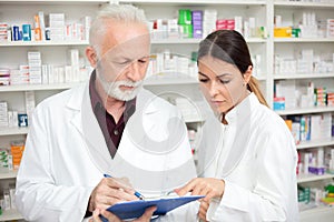 Serious young female and senior male pharmacists working in a drugstore, man writing data to a clipboard