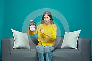 Serious young girl pointing on alarm clock, hinting at deadline, sitting on sofa. Time management, lack of time concept