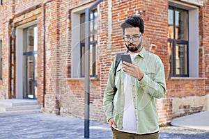 Serious young Indian man standing with backpack on city street, using mobile phone, reading and writing messages, making