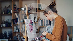 Serious young lady art student is painting flowers on canvas in studio holding brush and palette with paints. Lifestyle