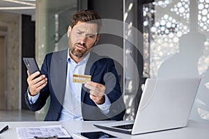Serious young male businessman sitting in office at desk with laptop and worriedly looking at credit card, holding phone