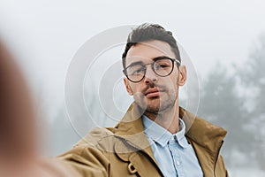 Serious young male is making self portrait on camera outdoors. Handsome traveler man wear coat, blue shirt and eyeglasses making