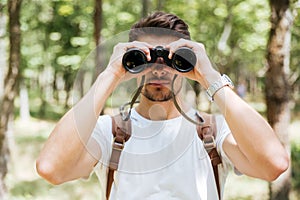 Serious young man with backpack using binoculars in forest