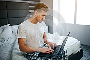 Serious young man in bed this morning. He work at home. Guy type on laptop keyboard and look at screen. Daylight.