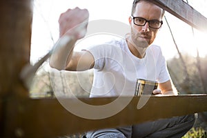 Serious young man in casual clothing painting wooden fence in his backyard
