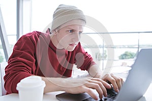Serious young man in casual clothing, sitting at the table and typing the text on the keyboard of the laptop