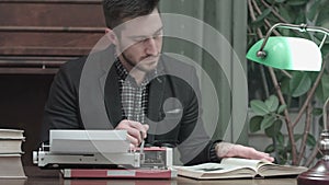 Serious young man with glasses sitting at the desk with typewriter and reading book