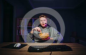 Serious young man in a headset sits at night at the computer and eats chips from a plate, looking intently at the camera