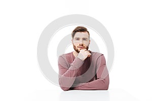 Serious young man looking at the camera sitting at the table isolated. Thinking pose with hand on the chin