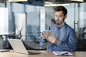 A serious young man is sitting in a modern office in a blue shirt at his desk and using a tablet