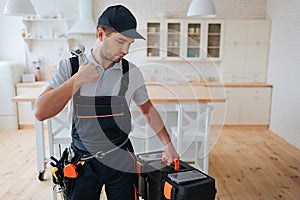 Serious young man stand in kitchen and look at tool box in his hand. Instruments on belt. Concentrated guy in uniform.