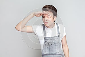 Serious young man standing keeps hand near forehead, looks far away searches something on horizon.