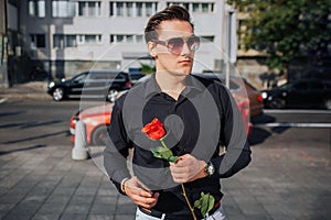 Serious young man wears sunglasses and looks to right. He hold red rose and phone in hands. He wait. It is sunny outside.