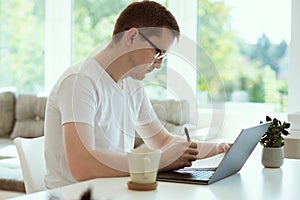 Serious young man working online with laptop at home, holding paper sitting at office desk