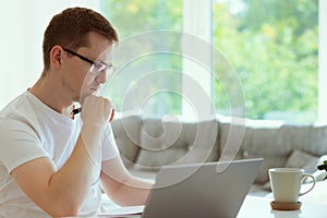 Serious young man working online with laptop at home, holding paper sitting at office desk