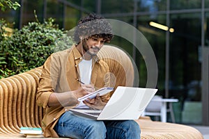 Serious young Muslim man sitting outside on a bench wearing headphones and using a laptop, writing in a notebook