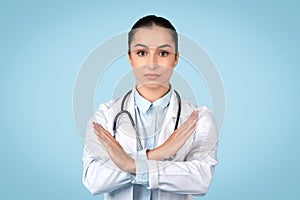 Serious young woman doctor in white coat making X sign with her arms, signaling denial or prohibition, blue background