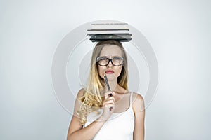 Serious young woman in glasses holding pen in her hand and books on her head  white background