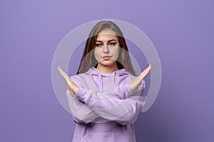 Serious young woman with long hair shows cross stop gesture, looking unamused, saying no, forbid something, wears purple hoodie,