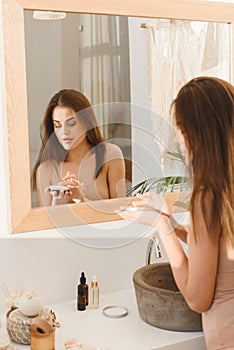 Serious young woman moisturizes face skin with cream in the morning in the bathroom. The concept of clean skin, facial
