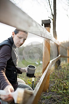 Serious young woman repairing and painting wooden fence