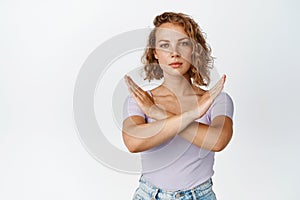 Serious young woman shows cross stop gesture, looking unamused, saying no, forbid something, standing over white photo