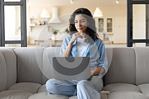 Serious young woman sitting on couch with laptop computer, working or studying from home