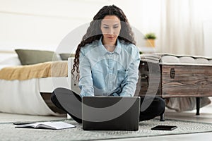 Serious young woman using laptop, having remote job, sitting on floor in bedroom, full length