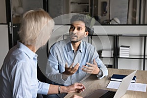 Serious younger Indian professional man talking to elder colleague woman
