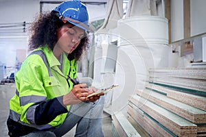 Seriously African woman worker with curly hair wearing helmet and safety vest, writing on file folder of document, inspecting wood