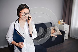 Serius worried young woman talk on phone. She stand in room. Her patient lying on couch behind. Small sick girl sleep. photo