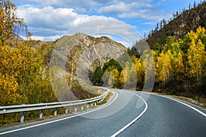 Serpentine asphalt road among high snow-capped mountain peaks, yellow desert, autumn green forest and blue sky. Car on