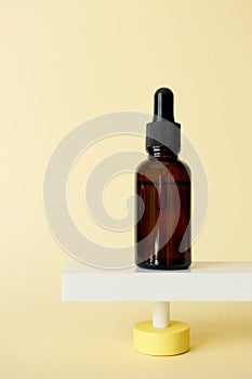 Serum facial moisturizer bottle standing on abstract pedestal on pastel yellow background with copy space, front view. Blank brown photo