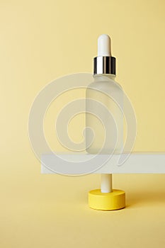 Serum facial moisturizer bottle standing on abstract pedestal on pastel yellow background with copy space, front view. Blank glass photo