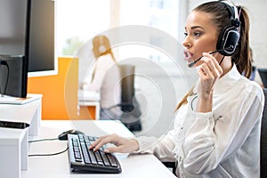 IT service desk female operator talking to client using headset and working on computer in call center office.