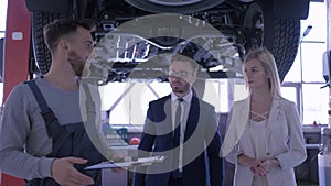 Service station, male master talking to a young couple near automobile raised on hydraulic lift gives keys and shakes