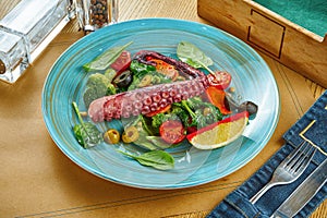 Serving of appetizing salad with octopus tentacle and steamed vegetables on a blue plate on a wooden background. Restaurant table