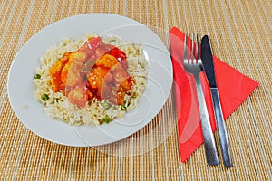 Serving of Chinese style sweet and sour chicken with rice on a white plate and bamboo table. Metal fork and knife on a red napkin