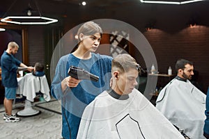 Serving client in barbershop. Professional young barber girl drying hair of a handsome guy, making trendy haircut. Focus