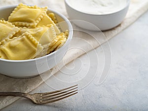 Serving of Italian ravioli, fork and sour cream sauce on a white plate. Pastel shades. There are no people in the photo. Close-up