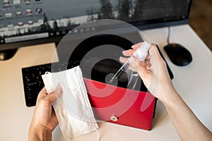 A set of anti-coronavirus antiseptic and medical mask. The girl puts the antiseptic in a red bag. Desktop with computer, office