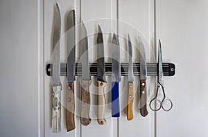 A set of chef's knives with different handles and different lengths hang on a magnetic pad on a white wall plus scissors