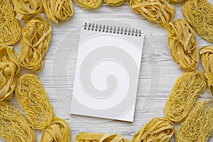 Set of different uncooked pasta with blank notepad on white wooden background, top view.