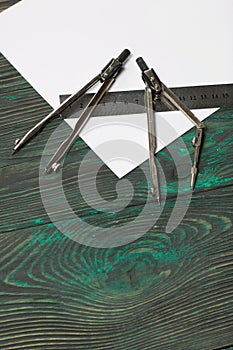 Set for drawing. Two compasses, a metal ruler and a sheet of white paper. They lie on pine boards painted in black and green